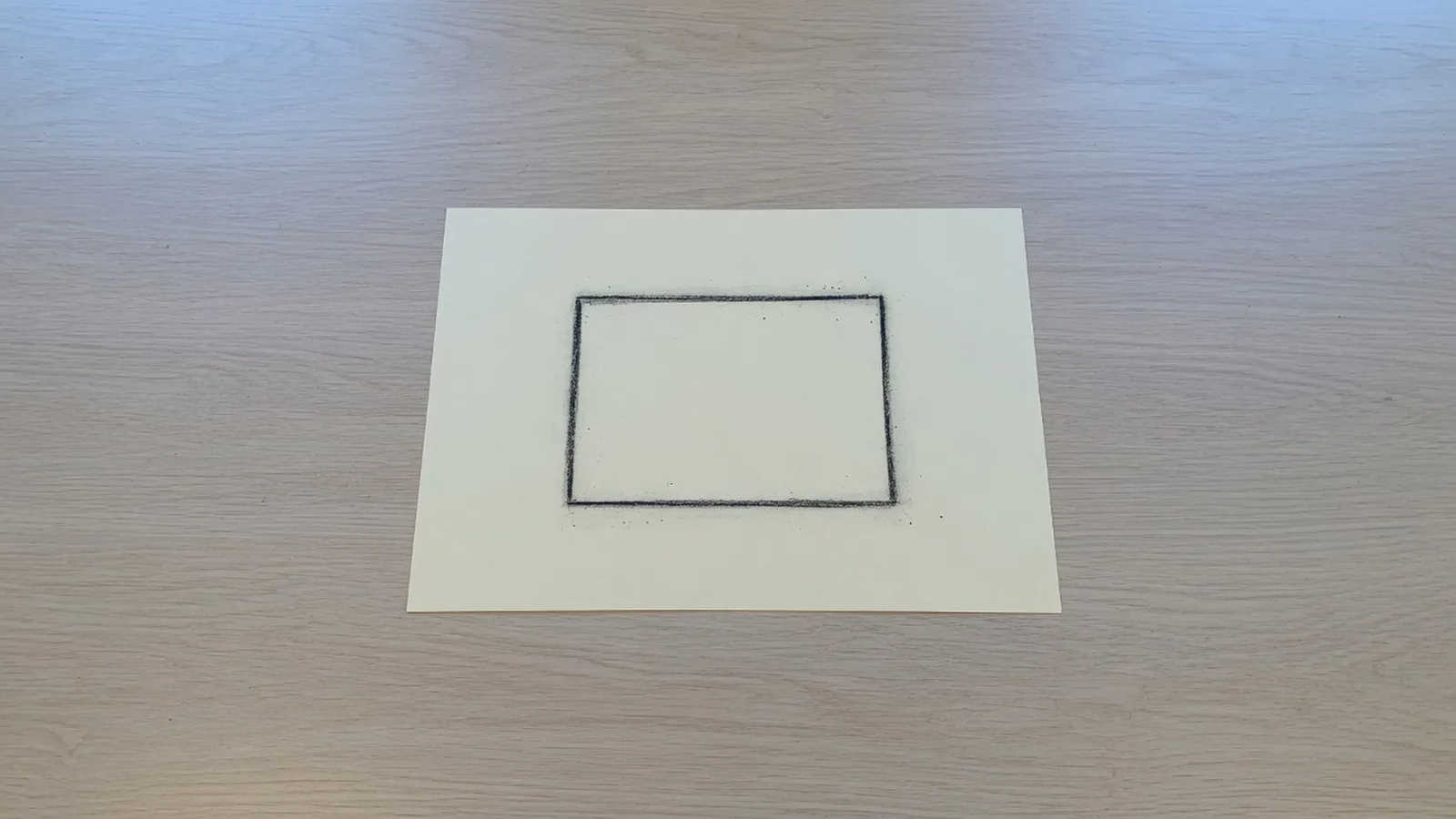 Vue de dessus : une seule feuille crème au centre d'une grande table de chêne clair, un rectangle au fusain tracé au milieu de la page