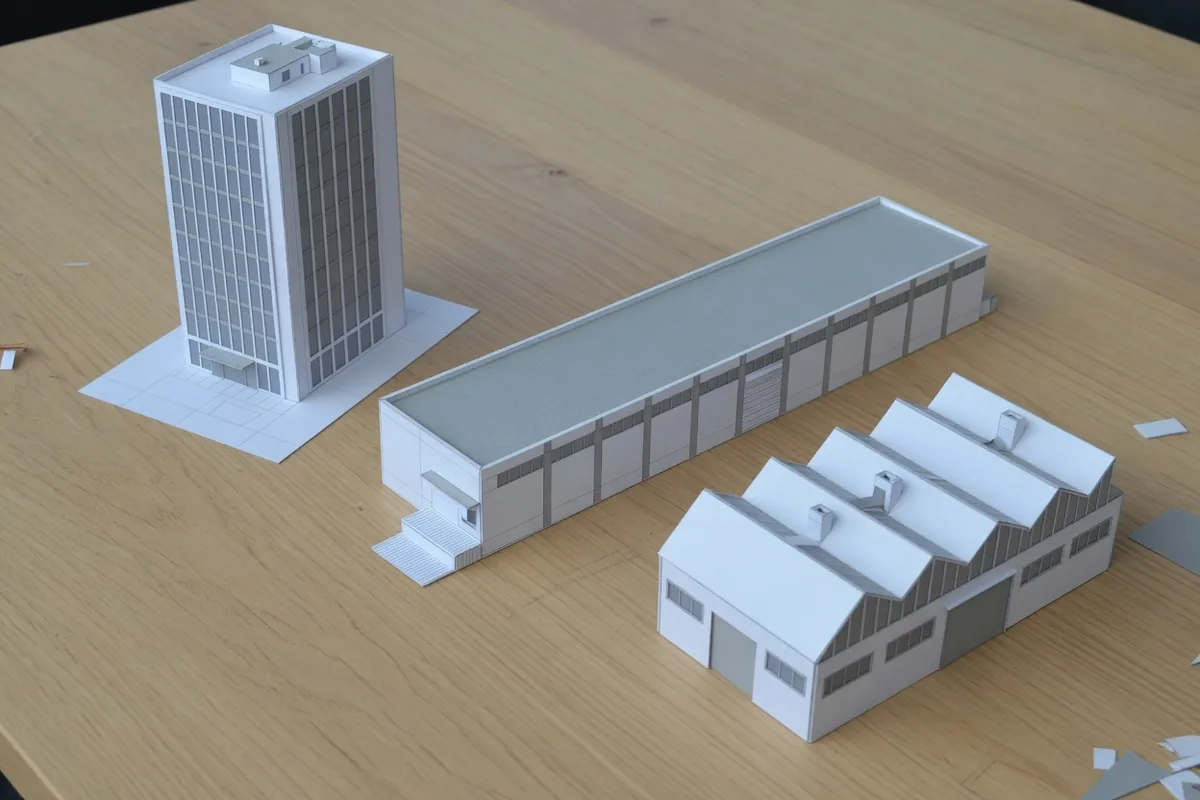 Trois maquettes blanches sur une table de chêne : une tour, un long bâtiment bas et un atelier à toits en dents de scie.