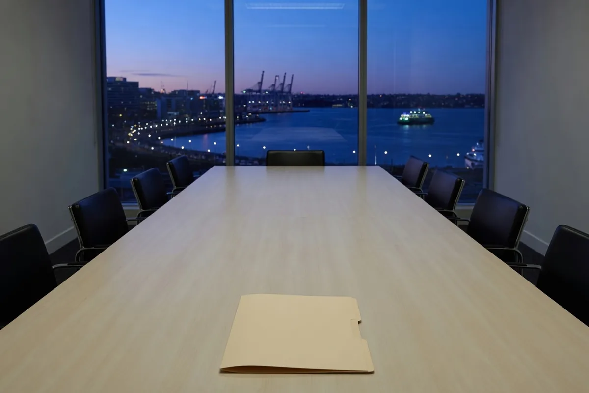 Une longue table de réunion vide au crépuscule, une chemise fermée au premier plan, un port éclairé derrière la vitre