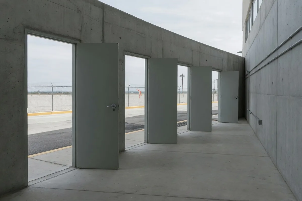 Quatre portes grises ouvertes dans un couloir de béton, donnant toutes sur le même extérieur pâle.